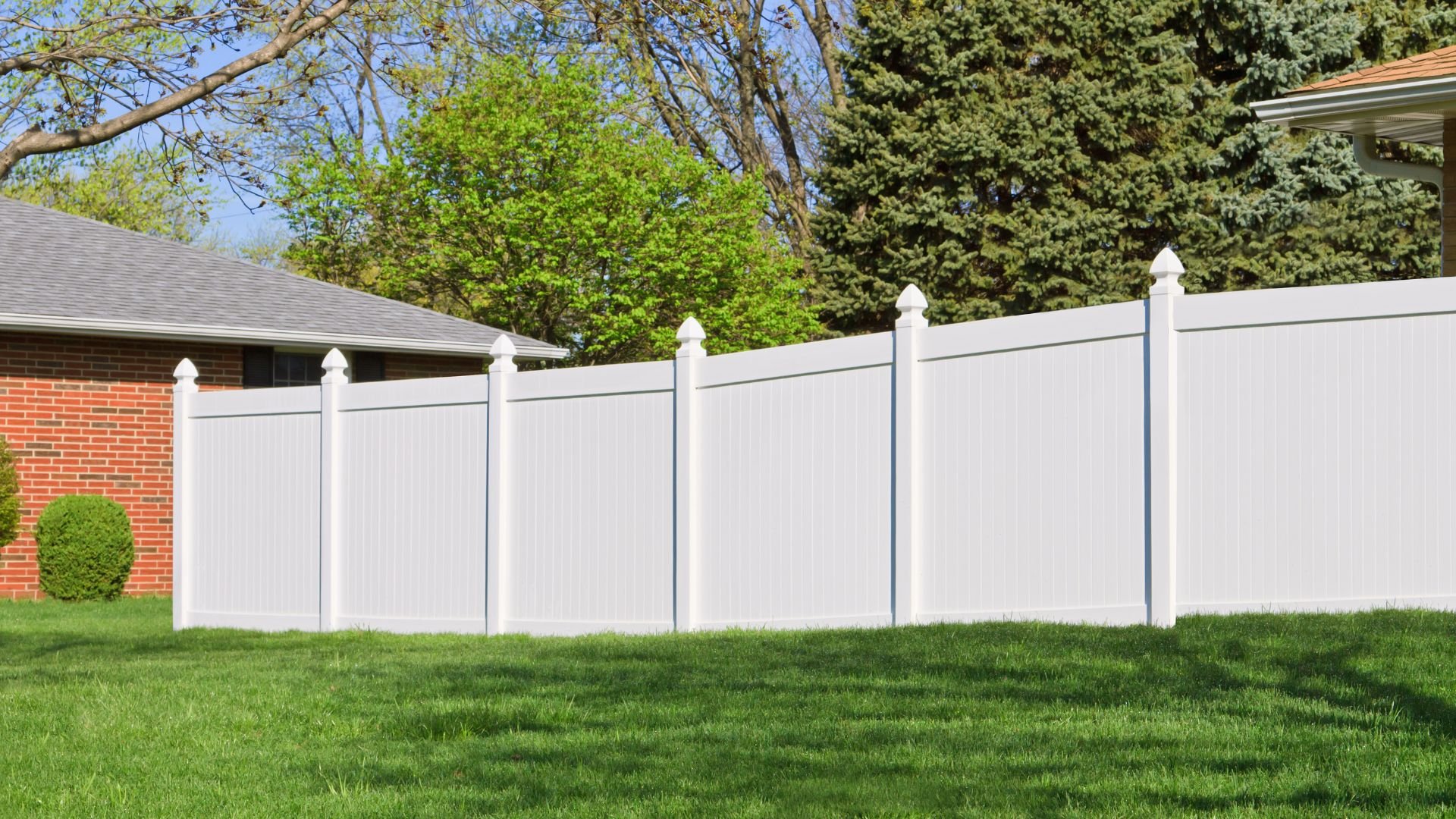 White vinyl fence surrounding a green lawn with trees and brick house
