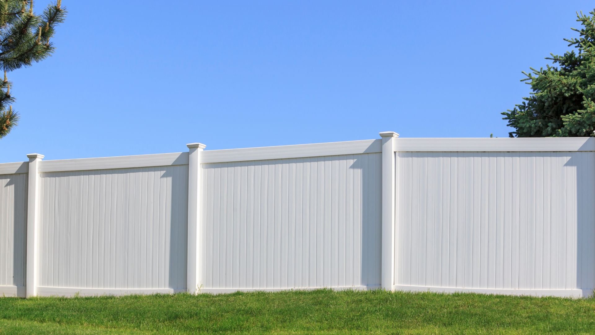 White vinyl fence with green grass and trees against blue sky