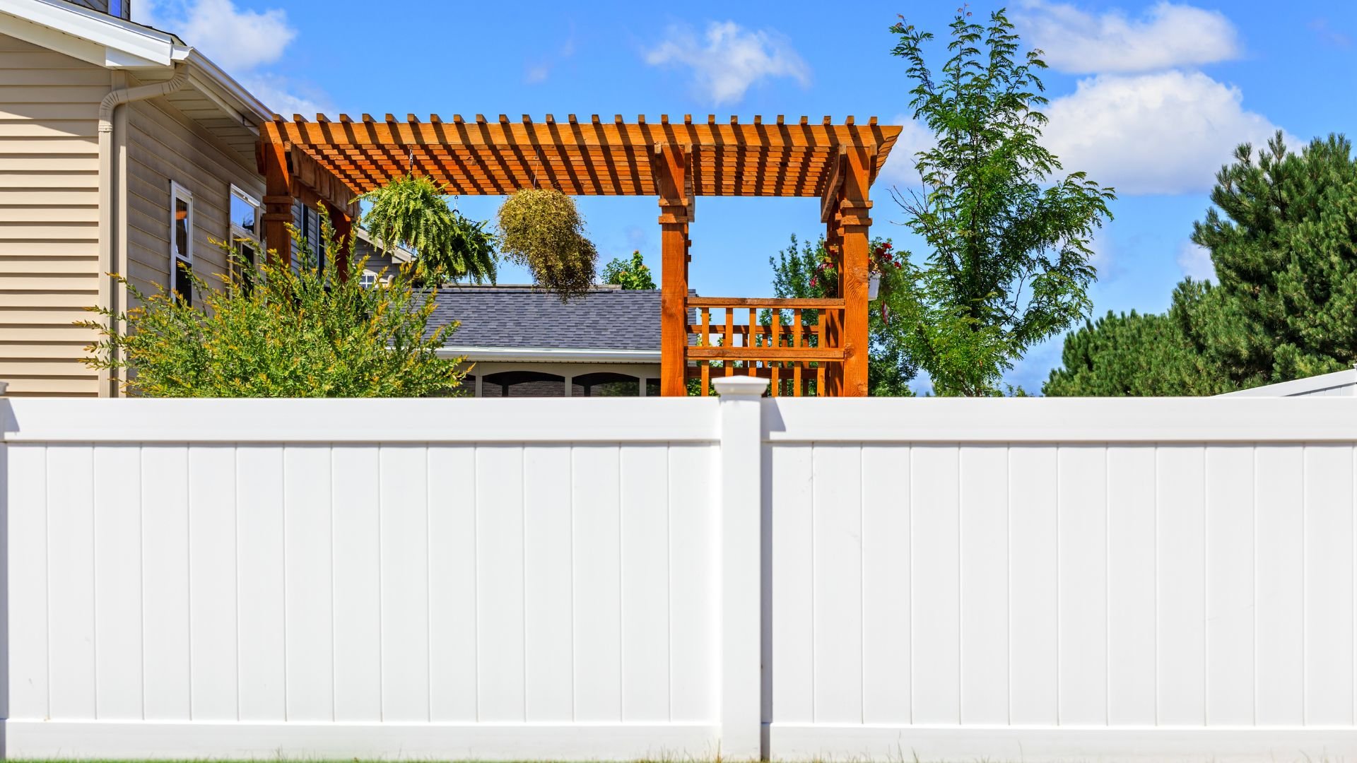 Wooden pergola with hanging plants behind a white vinyl fence